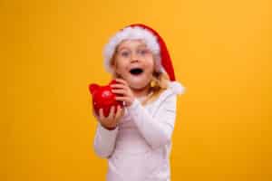 Little Blonde Girl In Santa Hat Holding Piggy Bank On Yellow Bac